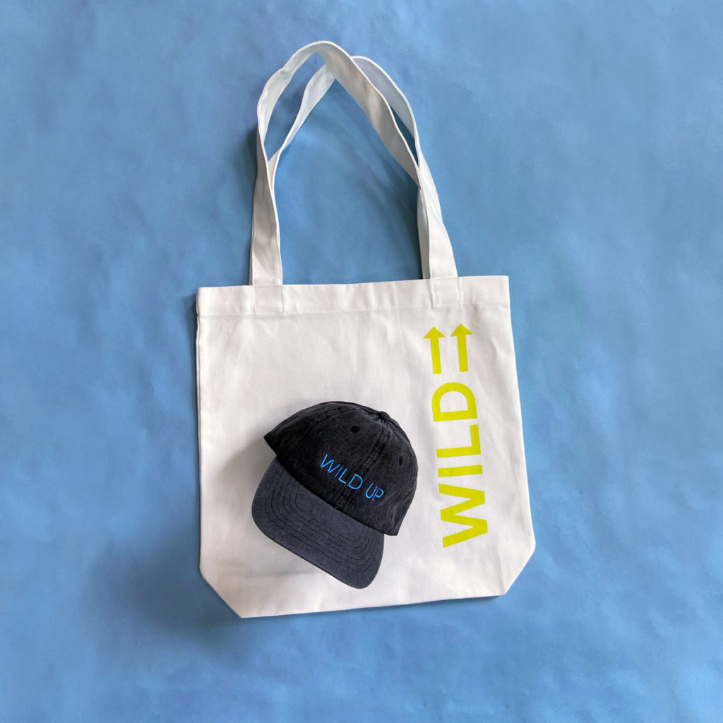 white tote bag with wild up logo in bright yellow green below one handle and a denim hat embroidered with the words "wild up"