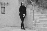 Person with long dark hair and glasses leans against a rough stucco wall beside a concrete staircase.
