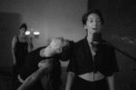 A woman sings into a microphone while another vocalist leans back dramatically toward her, in a dimly lit studio.