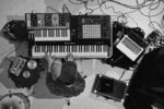 Overhead view of a musician playing a Rhodes keyboard surrounded by synthesizers, a laptop, mixer, and tangled cables.
